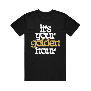 image of a black tee shirt on a transparent background. front has full body print in white that says it's your golden hour. the word golden is printed in gold shimmer ink